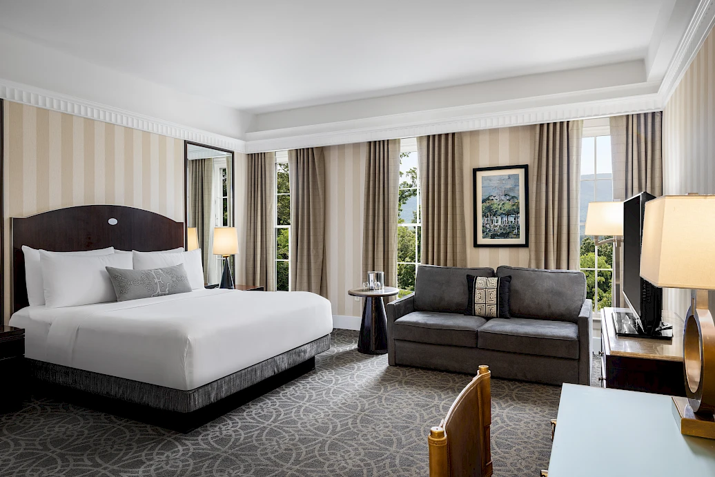 The image shows a modern hotel room with a large bed, a couch, a desk, and several lamps. There are large windows with curtains.