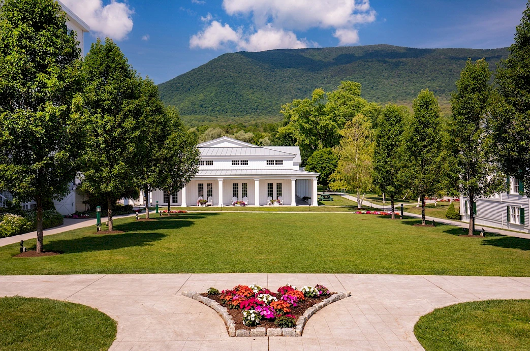 White building with columns, surrounded by greenery, trees, and a mountain in the background. Flower bed in the center foreground.