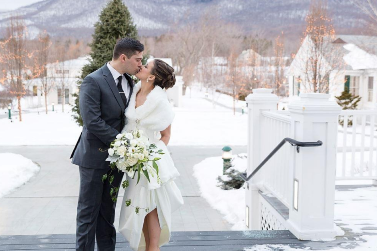 A couple dressed in wedding attire shares a kiss outdoors in a snowy landscape, with mountains and decorated trees in the background.