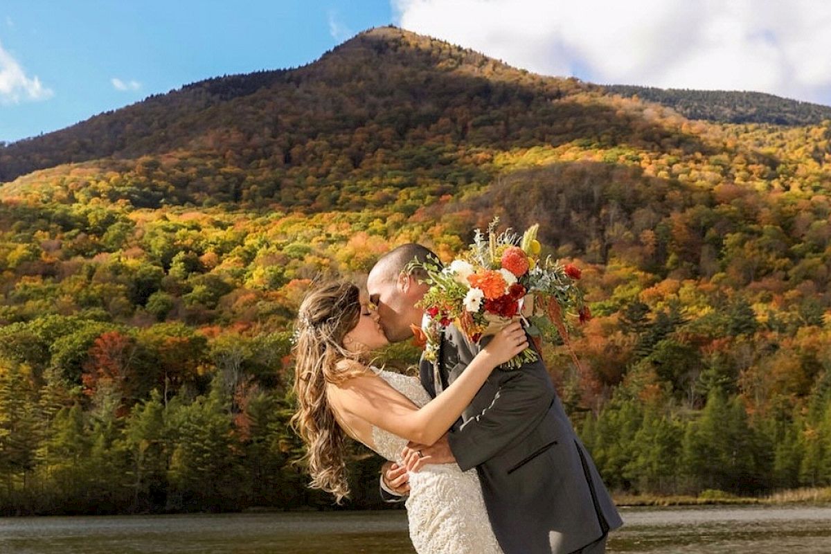 A couple shares a kiss in front of a picturesque mountain backdrop with autumn foliage, the bride holding a bouquet.