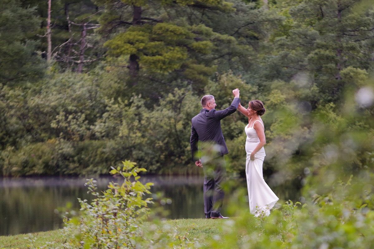 A couple in formal attire is dancing outdoors near a lake, surrounded by lush greenery, creating a romantic and serene atmosphere.