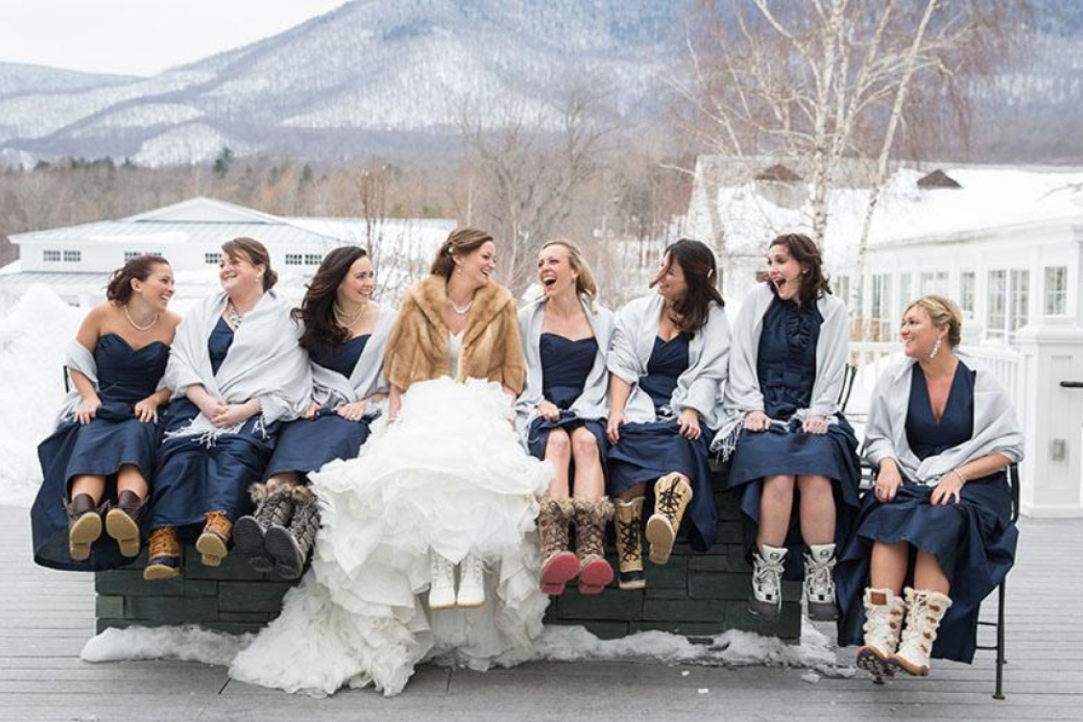 A bride and her bridesmaids sit on a bench in winter apparel and boots, with a snowy mountain landscape in the background.