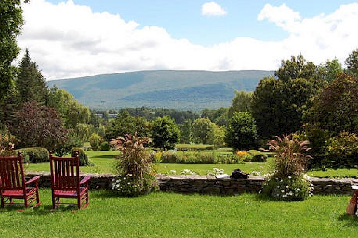 This image features a scenic landscape with wooden chairs on a grassy lawn, overlooking a garden and trees with mountains in the background.