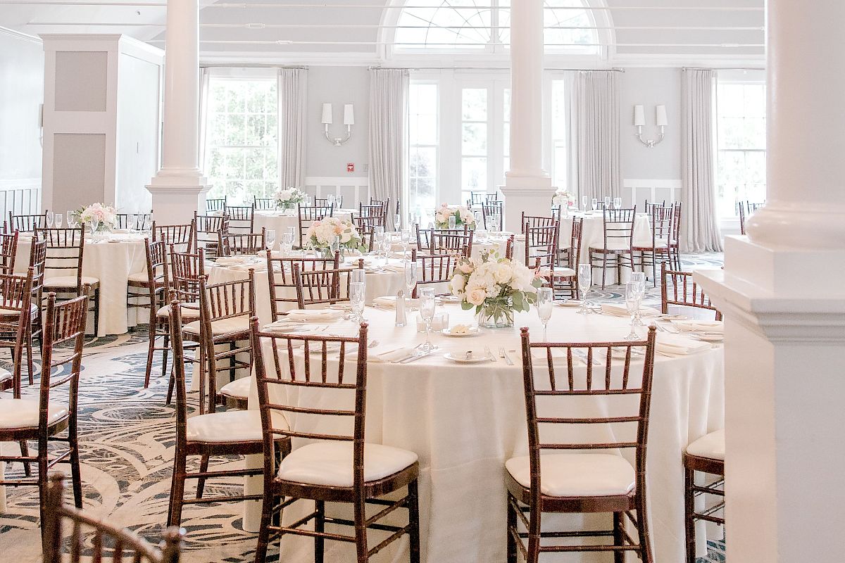 An elegant, light-filled banquet hall with white tablecloths, wooden chairs, floral centerpieces, and a chandelier overhead, ready for an event.