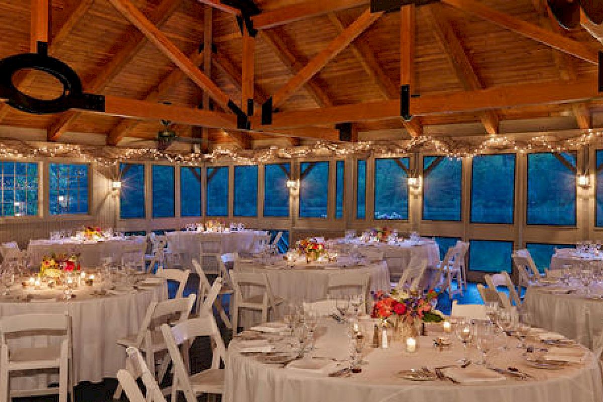 The image shows a decorated venue with round tables, white tablecloths, chairs, flower centerpieces, and string lights, likely set up for an event or reception.