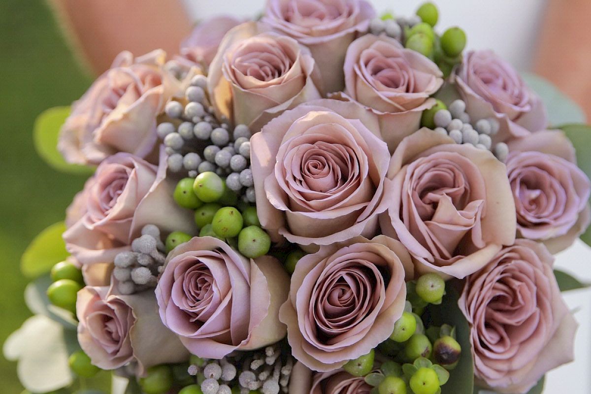 A close-up image of a bouquet featuring pale pink roses, small green berries, and other small, grayish-white flowers, being held by someone.