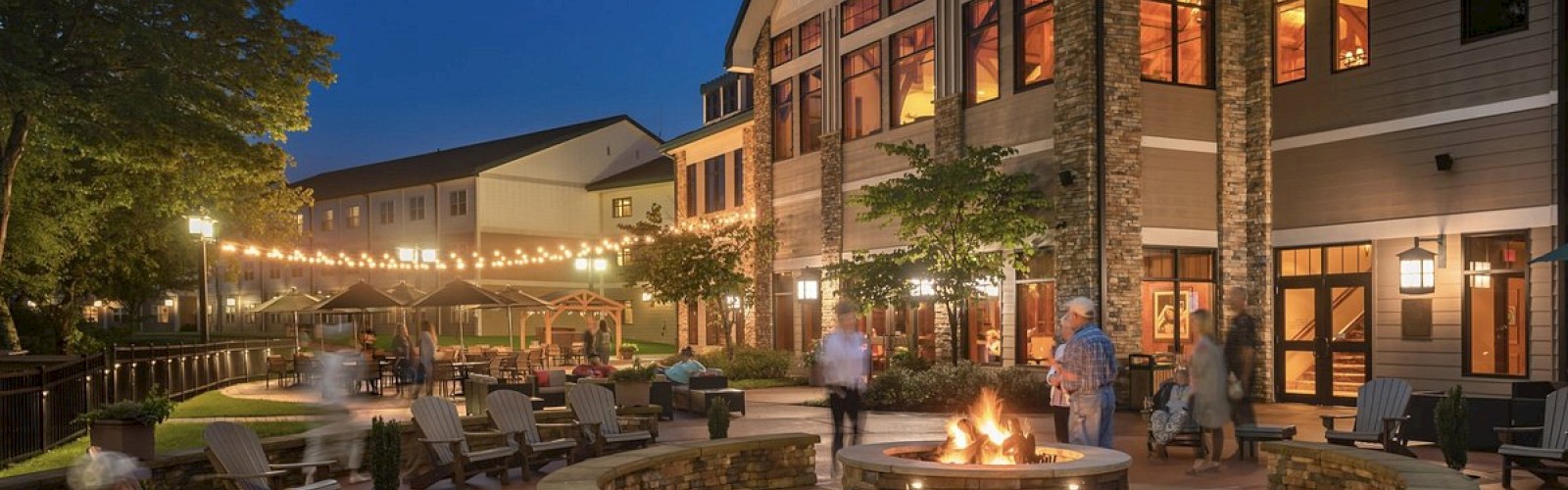 The image shows a cozy outdoor seating area with a fire pit, surrounded by a building, lights, and relaxed people in the evening.