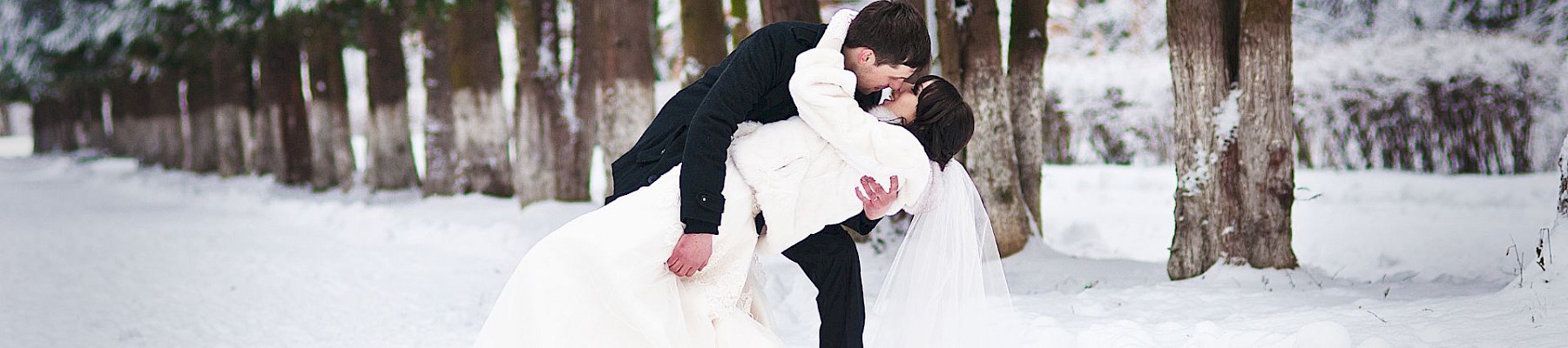 A couple in wedding attire sharing a kiss in a snowy forest with tall trees.
