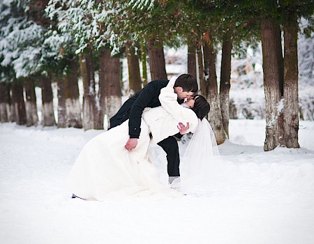 A couple in wedding attire sharing a kiss in a snowy forest with tall trees.