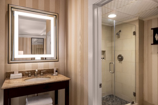 This image shows a bathroom with a lit square mirror above a marble countertop sink, and a glass shower with beige tiles.
