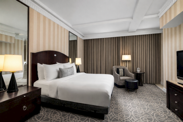A luxurious hotel room with a large bed, bedside lamps, a sitting area, TV, and elegant decor in neutral tones.