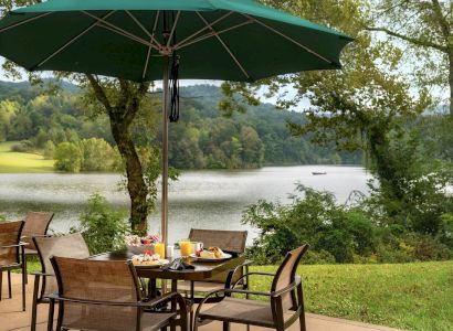 A scenic outdoor dining area with a table set for a meal under a green umbrella, overlooking a tranquil lake and lush greenery.