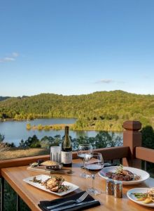 A table with food and wine is set on a balcony overlooking a scenic lake and forested hills under a clear blue sky.