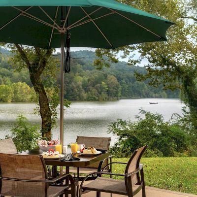 An outdoor dining setup with chairs, a table under an umbrella, food, and drinks, overlooks a scenic lake with trees in the background.