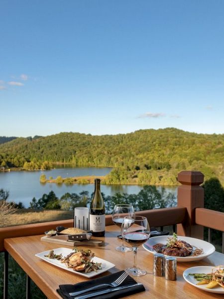 A table set with dishes and wine overlooks a scenic landscape with a lake, forest, and clear blue sky in the background.