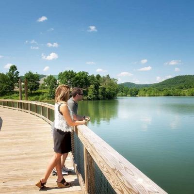 A couple stands on a wooden boardwalk overlooking a serene lake, surrounded by lush greenery and hills under a clear blue sky.