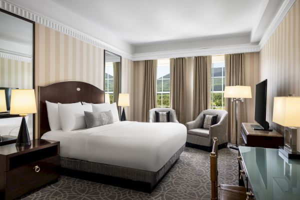 A hotel room with a large bed, two chairs, a TV, lamps on nightstands, a desk with a glass surface, and large windows with curtains.