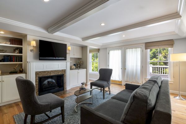 The image shows a modern living room with a gray couch, two chairs, a wall-mounted TV, bookshelves, a fireplace, and large windows with curtains.