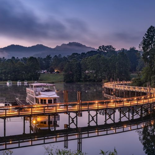 A serene landscape with a lit-up wooden walkway over water, a boat docked, surrounded by trees and mountains under a twilight sky.