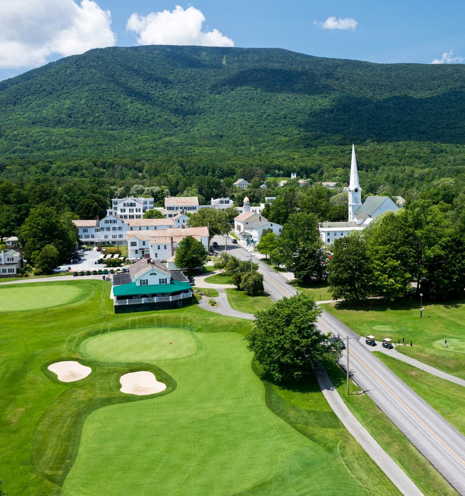 A scenic view of a small town with a church, surrounded by lush green golf courses and a mountain in the background on a sunny day.