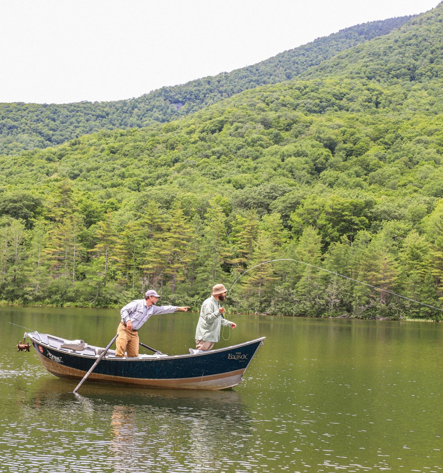 Two men are fishing from a boat on a calm lake surrounded by green, forested mountains.