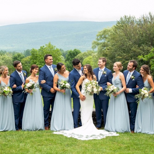 A wedding party is outdoors with a bride, groom, bridesmaids, and groomsmen posing on a green lawn with trees and mountains in the background.