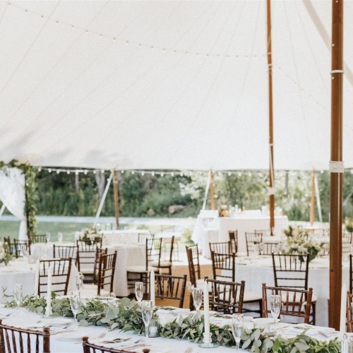 The image shows a decorated outdoor wedding reception under a large white tent with tables, chairs, floral arrangements, and greenery.