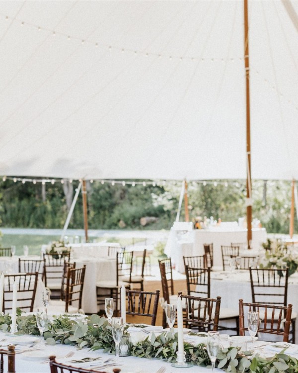 The image shows a decorated outdoor wedding reception under a large white tent with tables, chairs, floral arrangements, and greenery.