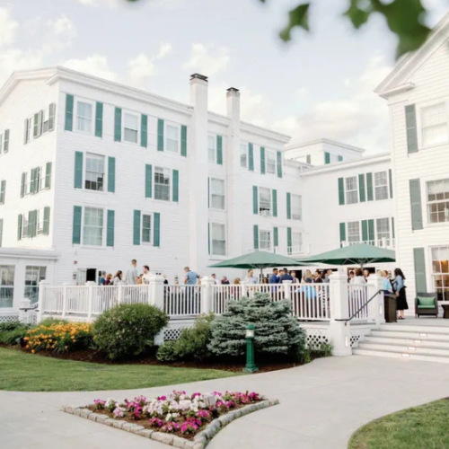 People gather for a social event on a white porch of a large, elegant white building with green shutters, surrounded by landscaped gardens.