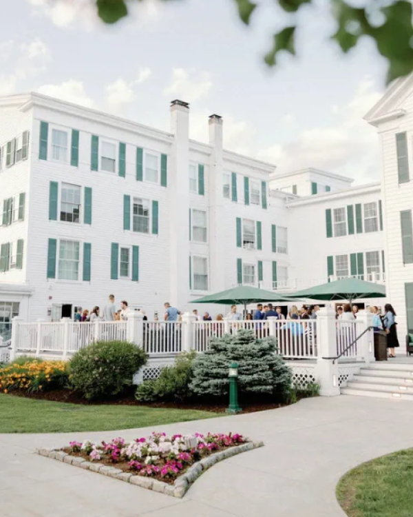 People gather for a social event on a white porch of a large, elegant white building with green shutters, surrounded by landscaped gardens.