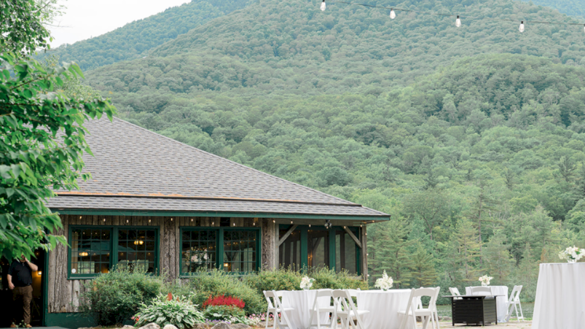 Outdoor event setup with decorated tables in front of a rustic building surrounded by lush green mountains.