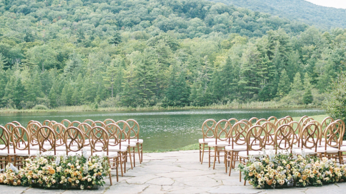 Chairs are set up outdoors near a lake, surrounded by lush green trees, indicating a scenic venue for a wedding or ceremony.