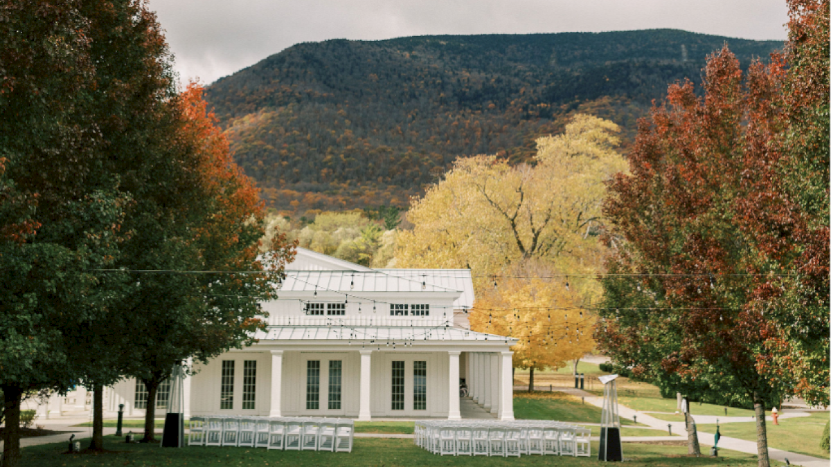 A white, classical-style building with columns stands outdoors, surrounded by trees with fall foliage and string lights overhead.