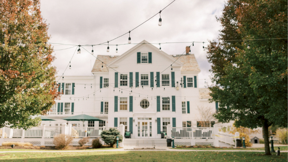 A large white house with green shutters, surrounded by trees and string lights, looks cozy and inviting for gatherings.