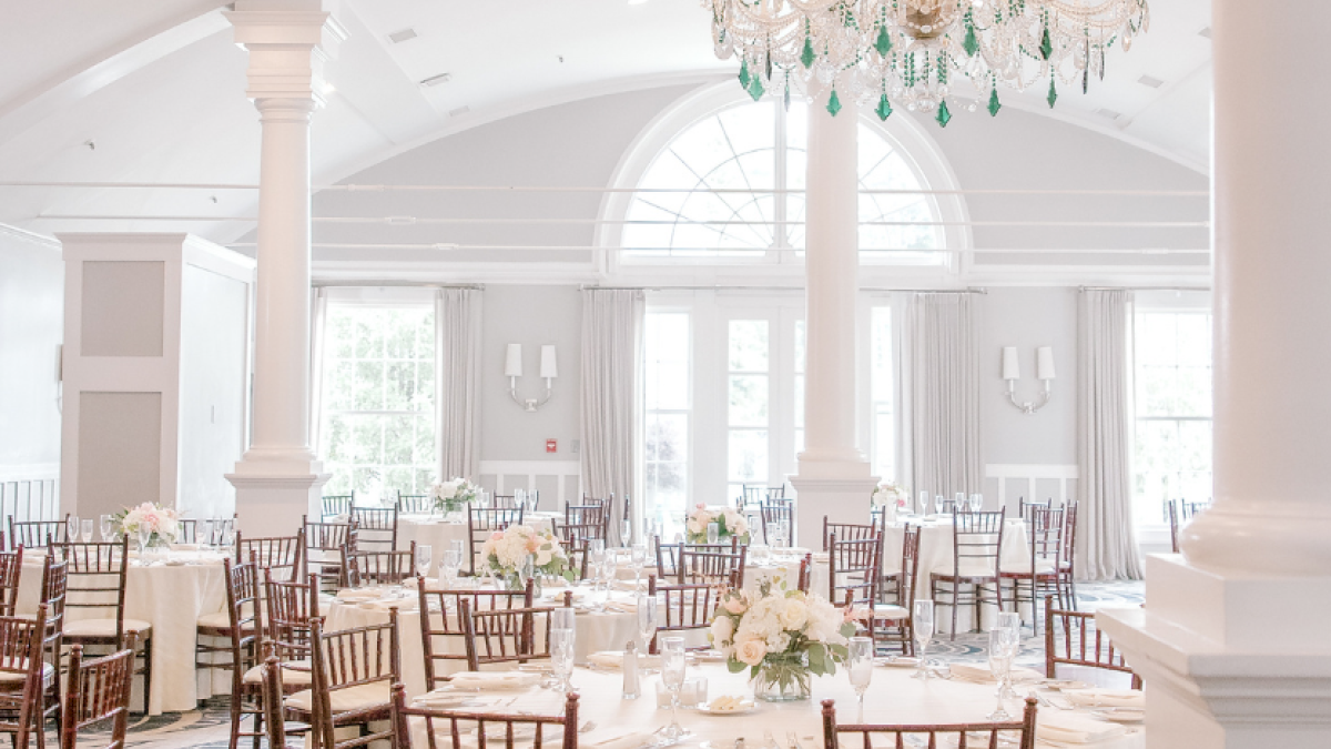 Elegant banquet setup with round tables, floral centerpieces, wooden chairs, and a large chandelier in a bright, airy room.