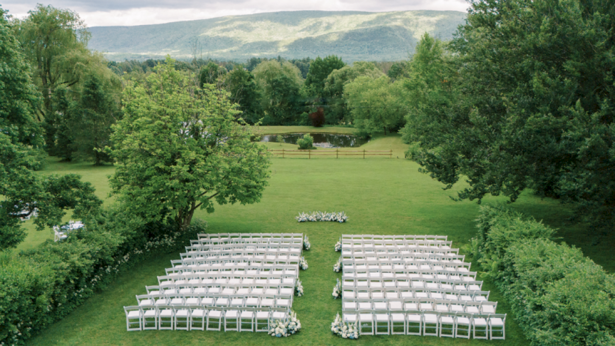 An outdoor wedding setup with white chairs, flowers, and a scenic green landscape with trees and a pond in the background.