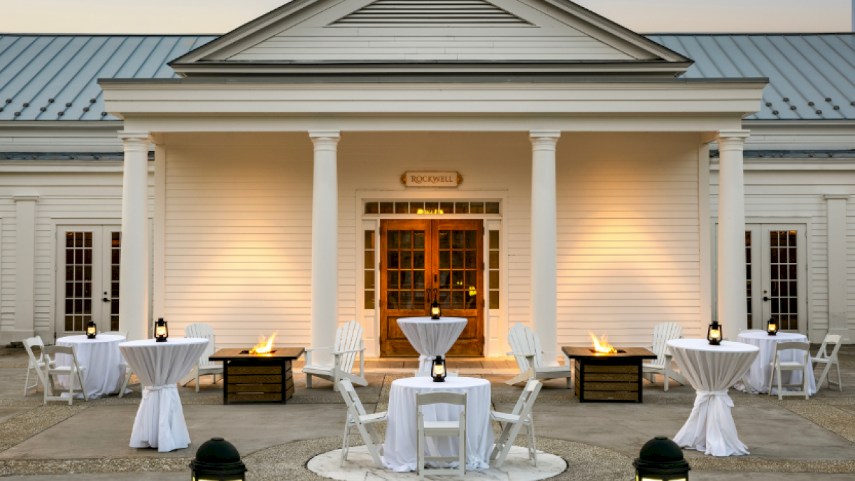 Elegant outdoor wedding setup with white tablecloths, lanterns, and fire pits in front of a classic, white building.