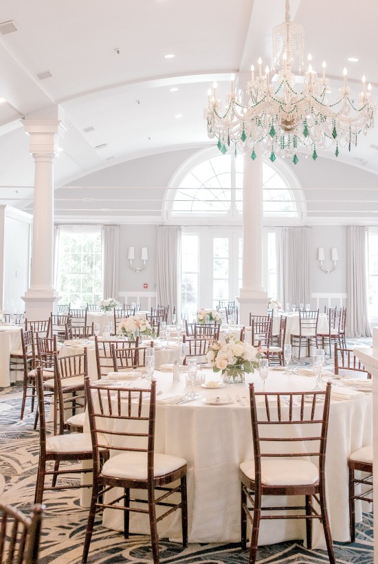 The image shows a beautifully decorated banquet hall with round tables, floral centerpieces, wooden chairs, large windows, and a chandelier.