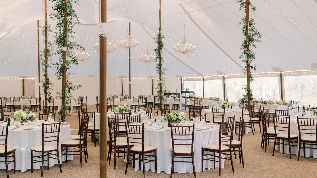 A spacious, elegant wedding reception under a large white tent, decorated with chandeliers, string lights, green foliage, and round tables.