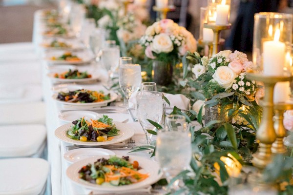 A beautifully decorated wedding reception table with floral centerpieces, candles, white tablecloths, and elegant place settings.