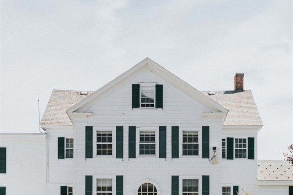 A large white house with black shutters, a circular window, and a welcoming front porch, surrounded by well-kept grass and plants.