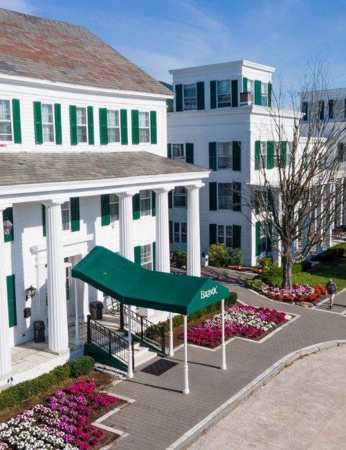 A large white building with green shutters, columns, and a green awning, surrounded by colorful flower beds and a few pedestrians.