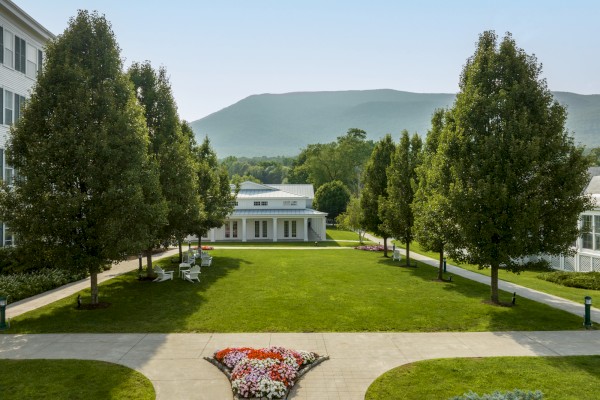 A peaceful park with trees, flower beds, benches, and a white building, with mountains in the background on a bright, sunny day.