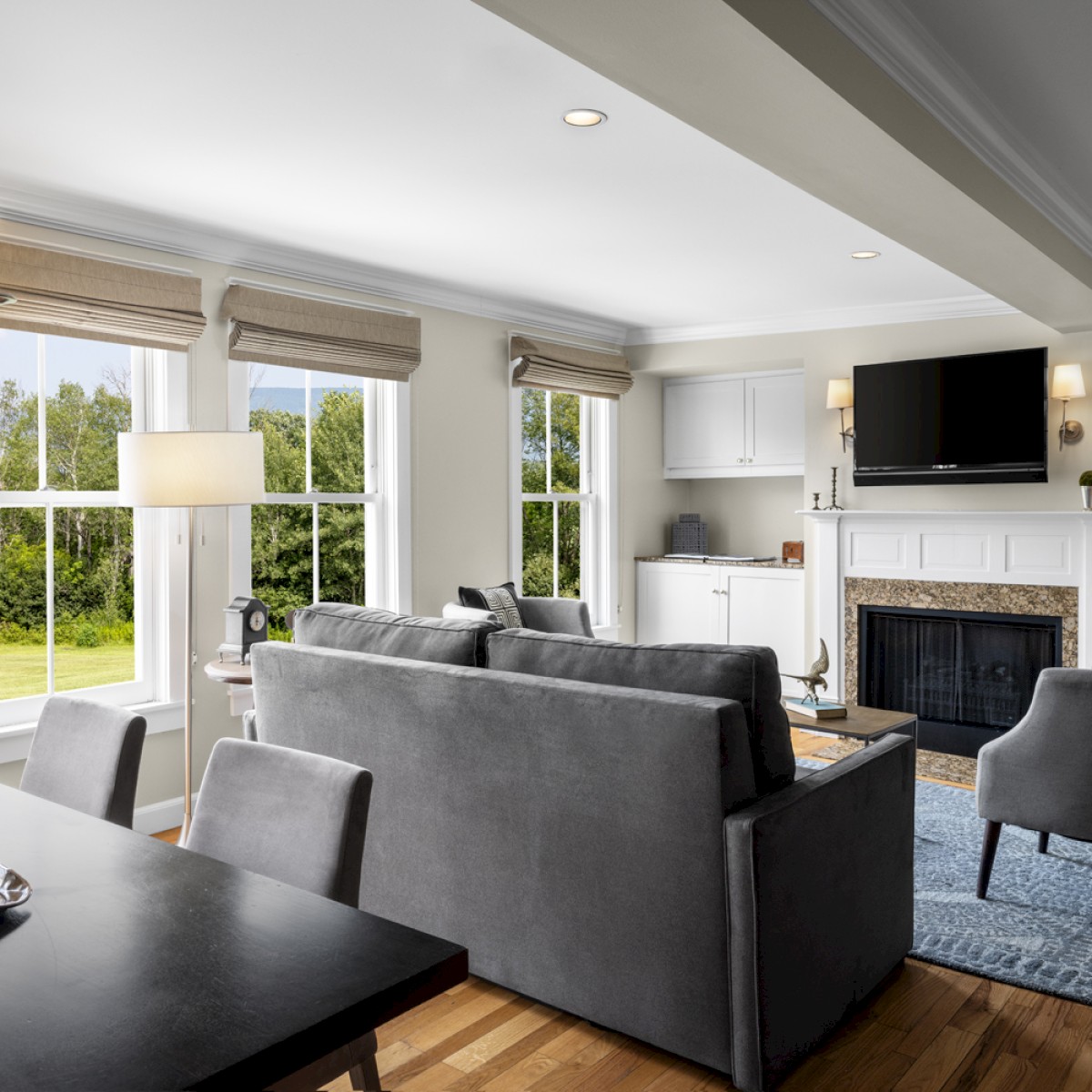 This image shows a cozy living room with a firelit fireplace, a large TV, gray sofas, a dining table, and natural light streaming in from large windows.