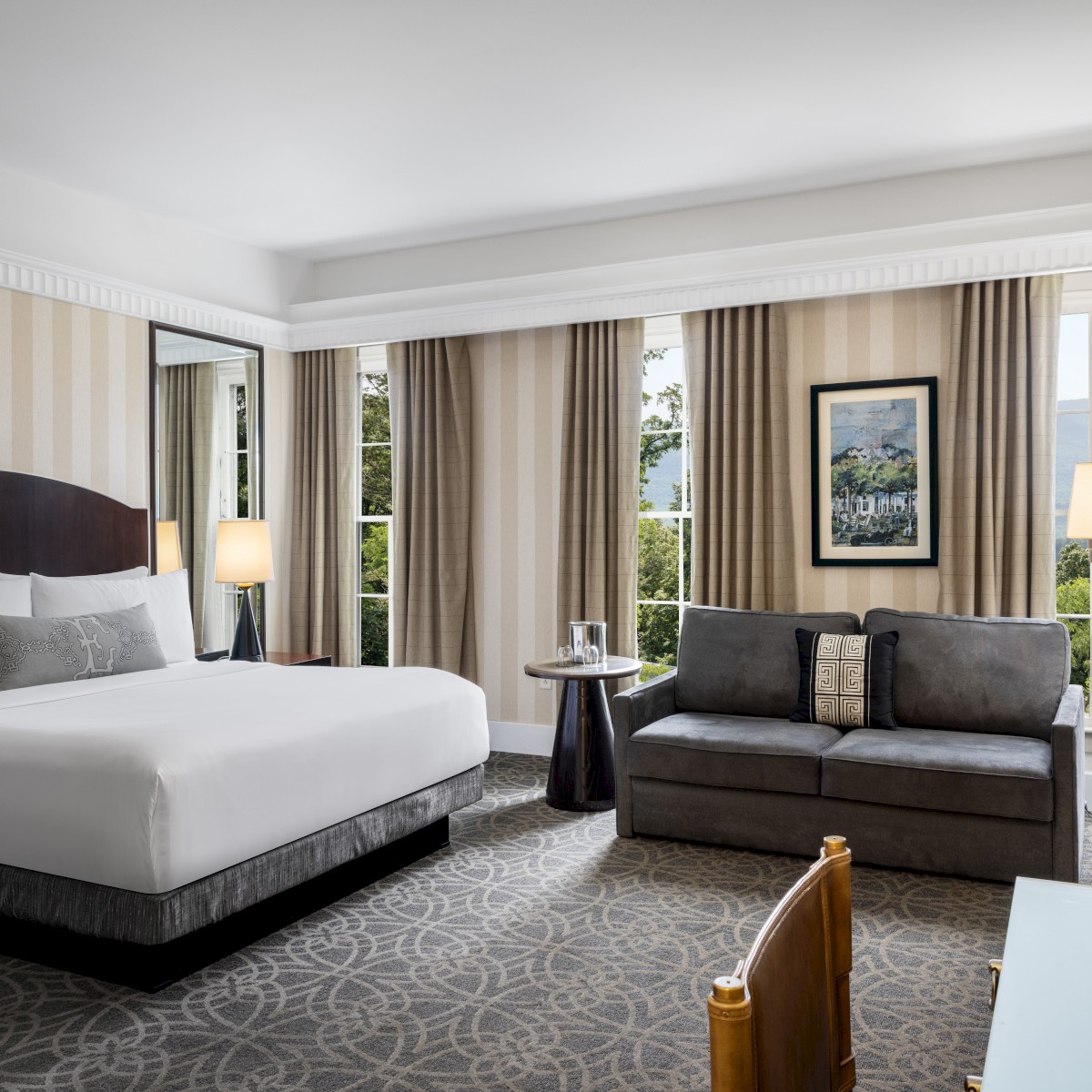 The image shows a modern hotel room with a large bed, a couch, a desk, and several lamps. There are large windows with curtains.