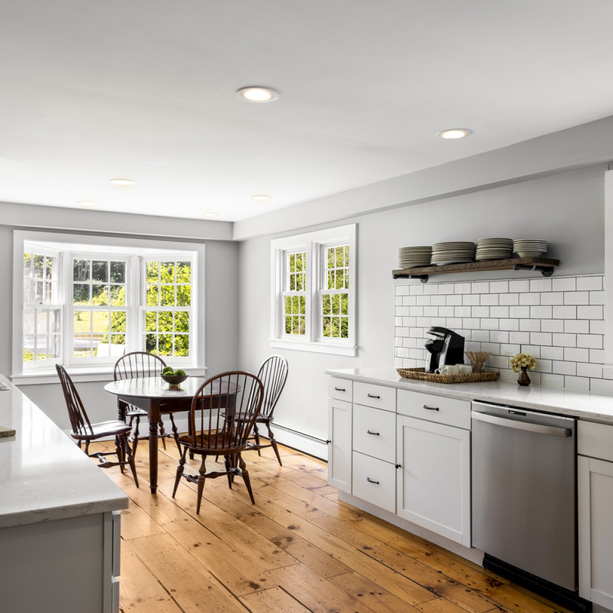 A modern kitchen with white cabinets, subway tile backsplash, wooden floors, open shelving, a dining table, and large windows providing natural light.