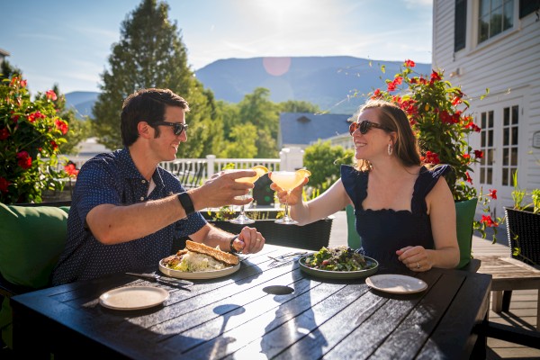 A man and woman happily toast cocktails outdoors on a sunny patio with food, flowers, and mountains in the background.