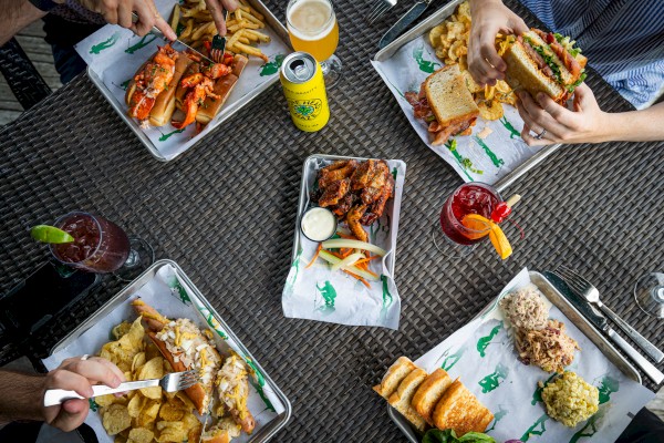 People enjoying a variety of food and drinks at an outdoor table, including sandwiches, fries, wings, and cocktails.