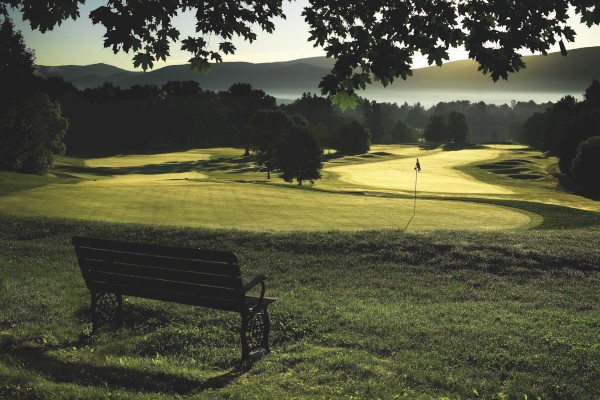 A peaceful golf course scene during early morning or late afternoon, with a bench under a tree overlooking the greens and mountains in the background.