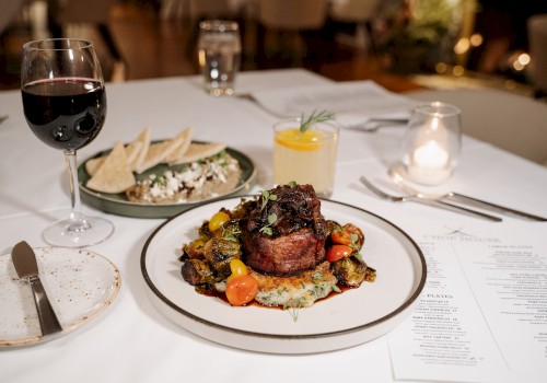 A plated steak with roasted vegetables, a glass of red wine, and other dishes on a dinner table set for a meal.